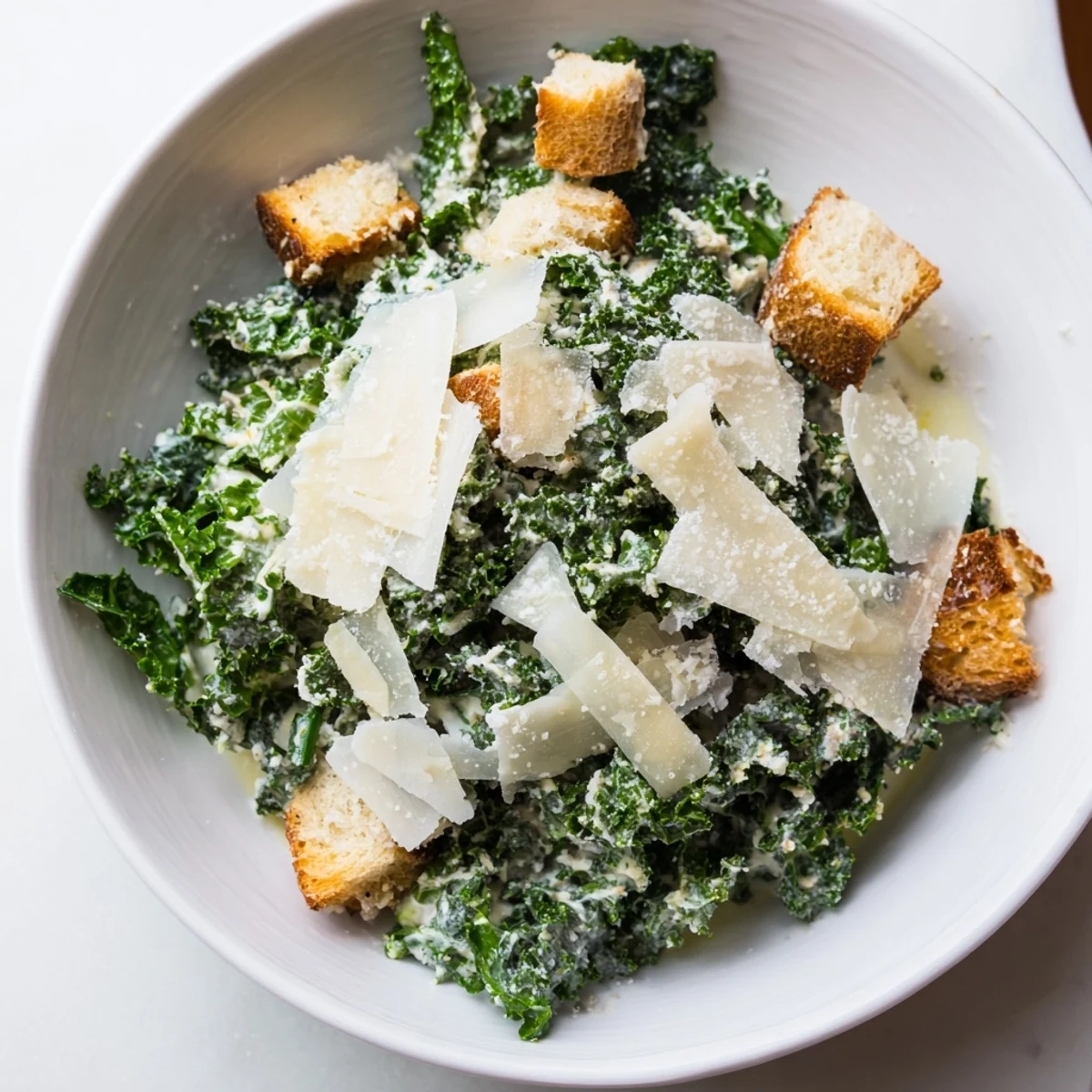 A close-up of a vibrant Kale Caesar Salad with shaved Parmesan and golden croutons on top.