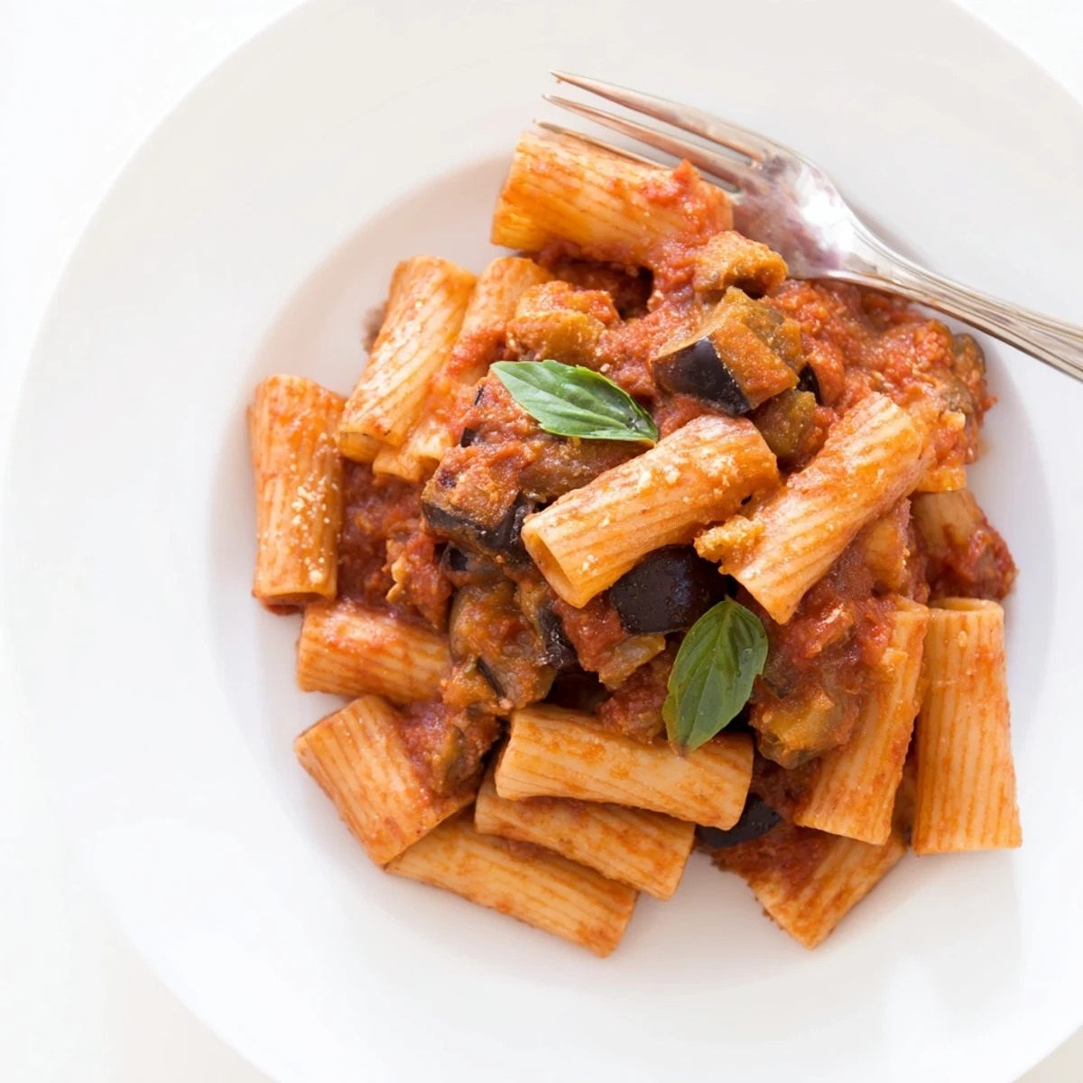 A close-up of Pasta Alla Norma highlighting golden roasted eggplant pieces, vibrant red tomato sauce, and a dusting of grated salty ricotta salata over al dente pasta.