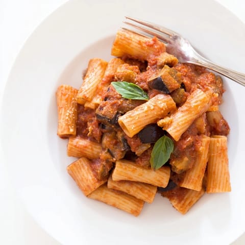A close-up of Pasta Alla Norma highlighting golden roasted eggplant pieces, vibrant red tomato sauce, and a dusting of grated salty ricotta salata over al dente pasta.